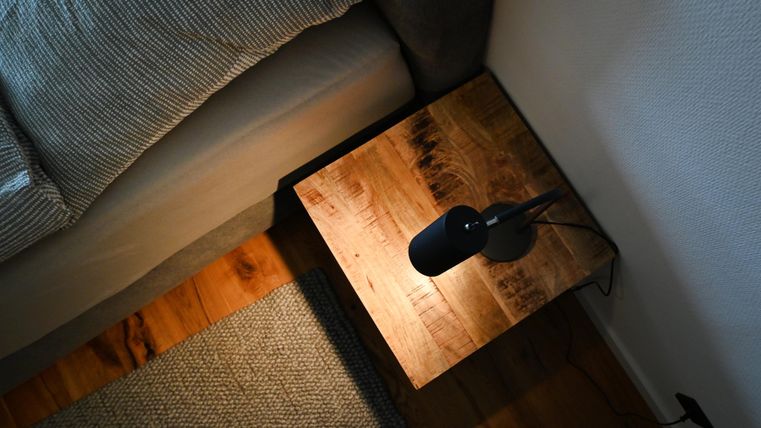 Ein Holztisch mit einer Lampe steht neben einem Sofa. Der Boden ist aus Holz, und ein Teppich liegt daneben.