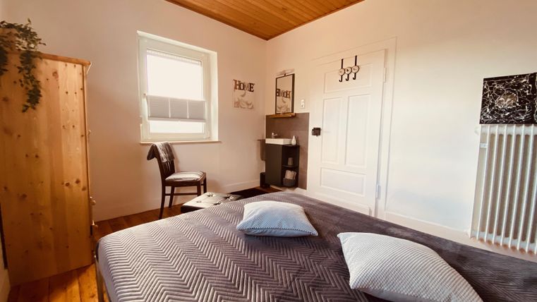 Ein gemütliches Schlafzimmer mit einem großen Bett und zwei Kissen. Es gibt einen Holzschrank, einen Stuhl und ein Fenster, das viel Tageslicht hereinlässt.