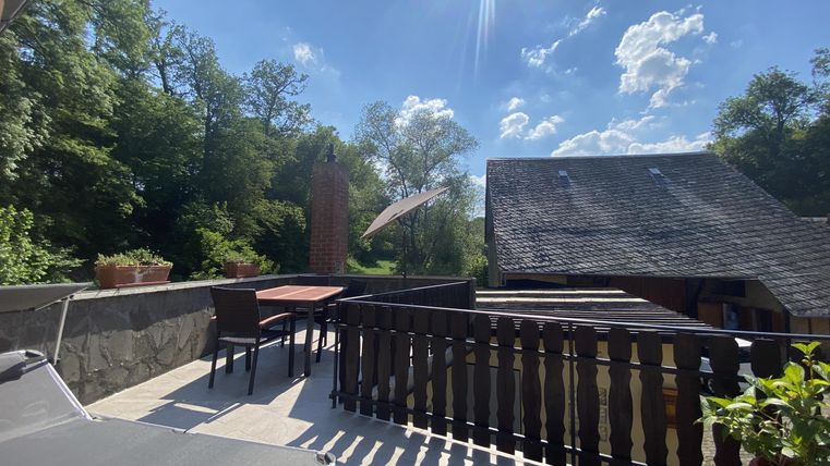 Eine Terrasse mit einem Tisch und Stühlen, umgeben von Bäumen. Der Himmel ist klar und sonnig.