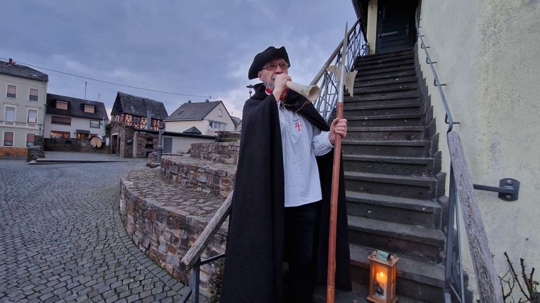 Een man in een mantel staat met een stok en een lantaarn op een trap. Op de achtergrond zijn oude huizen en een geplaveid plein te zien.