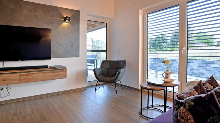 Ein modernes Wohnzimmer mit einem Fernseher an der Wand und einem bequemen Sessel. Große Fenster sorgen für viel Tageslicht und einen Blick nach draußen.