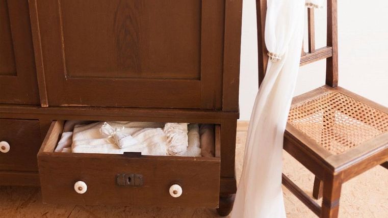 Une armoire brune avec un tiroir ouvert, dans lequel se trouvent des textiles blancs. À côté de l'armoire se trouve une chaise en bois avec un tissu blanc.