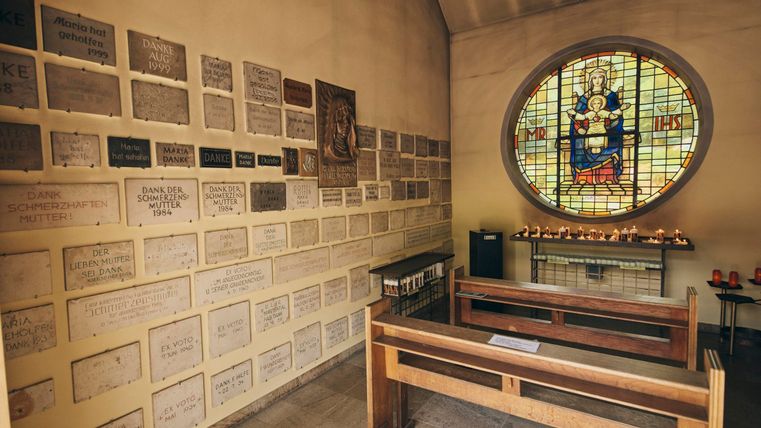 Eine ruhige Kapelle mit einer bunten Glasmalerei und einer Wand voller Gedenktafeln. In der Mitte stehen einfache Holzbänke für Besucher.