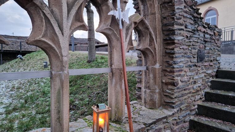 Ein historischer Steinbogen mit einer Laterne und einem langen Stock. Im Hintergrund sind grüne Wiesen und ein grauer Himmel sichtbar.