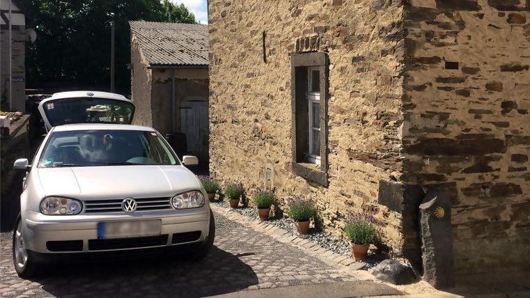 Ein grauer Volkswagen steht vor einem steinernen Haus. Im Hintergrund sind Gartenpflanzen und ein klarer Himmel zu sehen.
