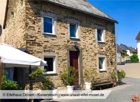 Ein gemütliches Steinhaus mit einem roten Eingang und großen Fenstern. Vor dem Haus gibt es einen kleinen Garten mit Pflanzen und einem Sonnenschirm.
