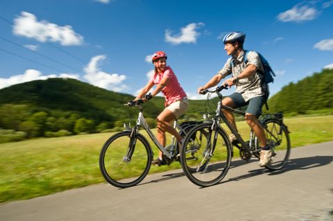 Ein lächelndes Paar fährt bei Sonnenschein auf Fahrrädern durch eine grüne Wiesenlandschaft mit bewaldeten Hügeln im Hintergrund und blauem Himmel mit weißen Wolken.