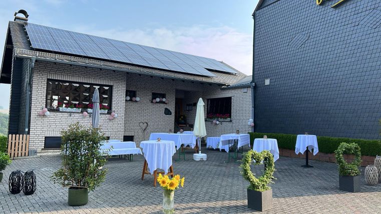 Een restaurant met een terras en witte tafels op een geplaveid plein. Op de achtergrond is een gebouw met zonnepanelen en een groene heg te zien.