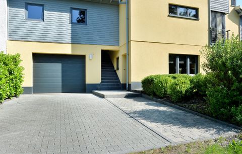Ein modernes Haus mit einem gelben Außenanstrich und einer gepflasterten Einfahrt. Grünes Gebüsch umgibt den Eingang und die Garagentür.
