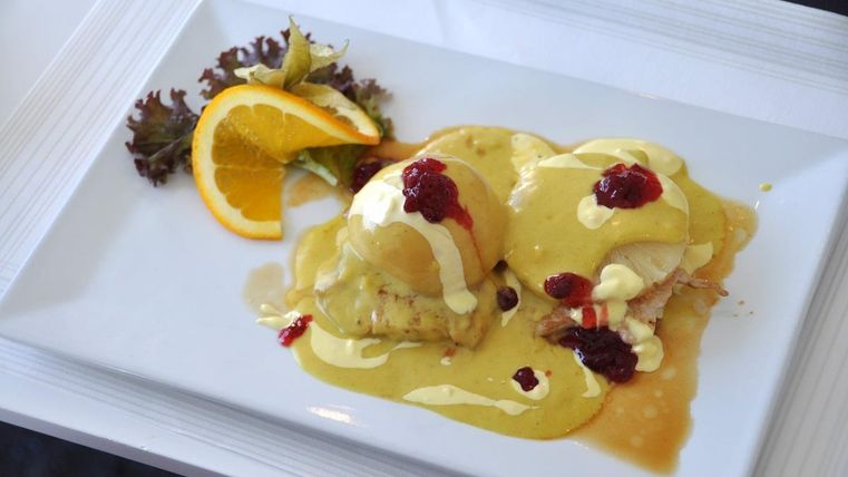 Ein Teller mit pochierten Eiern, garniert mit einer cremigen Sauce und Himbeermarmelade. Daneben befinden sich eine Orangenscheibe und etwas grüne Salatbeilage.