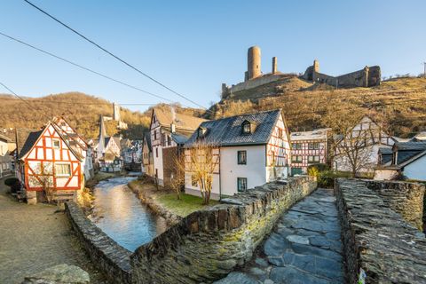 Eine malerische Landschaft mit charmanten Fachwerkhäusern und einem kleinen Fluss. Im Hintergrund erhebt sich eine beeindruckende Burg auf einem Hügel.