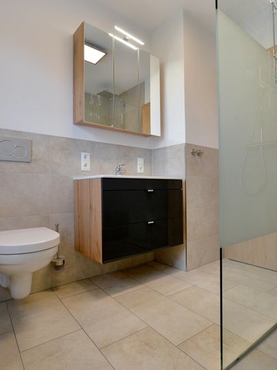 Ein modernes Badezimmer mit einem Wandwaschbecken aus Holz und Schwarz. Die Dusche ist durch eine Glaswand abgetrennt und der Raum wirkt hell und sauber.