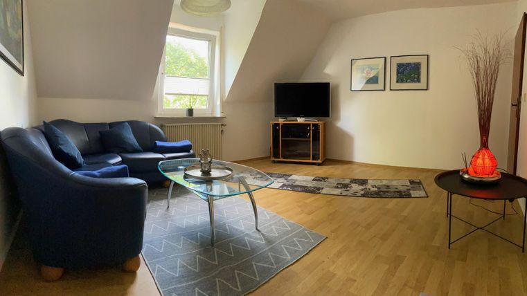 Ein gemütliches Wohnzimmer mit blauen Sofas und einem Glastisch. An der Wand hängen einige Bilder, und ein Fernseher steht in der Ecke des Raumes.