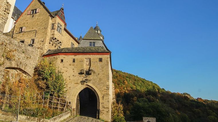 Ein historisches Schloss mit einer beeindruckenden Eingangstür. Die umliegende Landschaft ist mit Bäumen im Herbstlaub geschmückt.