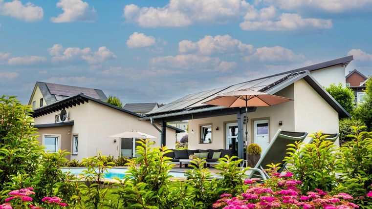 Ein modernes Haus mit einem Swimmingpool und einem schönen Garten. Bunte Blumen umgeben die Terrasse unter einem hellblauen Himmel.