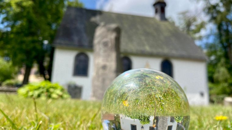 Eine kleine Kirche steht im Hintergrund, während eine Kristallkugel im Vordergrund die Szene reflektiert. Der Himmel ist blau mit einigen Wolken, und grünes Gras umgibt die Kirche.