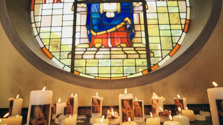 Eine Gruppe von brennenden Kerzen steht vor einem bunten Kirchenfenster. Das Fenster zeigt eine religiöse Darstellung.