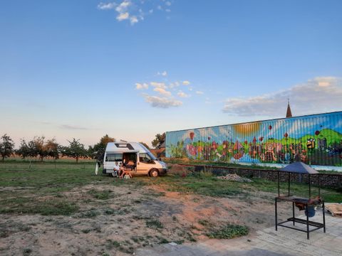 Ein Wohnmobil steht auf einem offenen Feld neben einer bunten Wandmalerei. Der Himmel ist klar mit einigen Wolken und es herrscht eine ruhige Atmosphäre.
