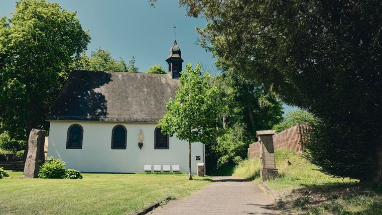 Eine kleine, malerische Kirche umgeben von Bäumen und grünem Rasen. Der Himmel ist klar und der Weg zur Kirche ist gepflastert.