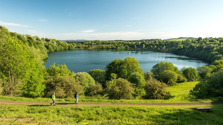 Eine malerische Landschaft mit einem ruhigen See und üppigem Grün. Zwei Personen spazieren entlang eines Weges am Ufer.