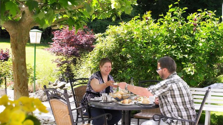 Eine schöne Außenterrasse mit einem Tisch, an dem zwei Personen eine Mahlzeit genießen. Die Umgebung ist grün und einladend, umgeben von Bäumen und Blumen.