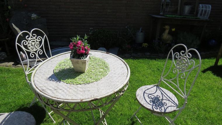 Ein kleiner Gartentisch mit zwei weißen Stühlen steht auf grünem Rasen. In der Mitte des Tisches befindet sich ein Blumentopf mit bunten Blumen.