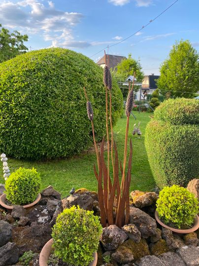 Een verzorgde tuin met ronde hagen en verschillende planten. Op de voorgrond staan metalen sculpturen die hoog uit de grond oprijzen.