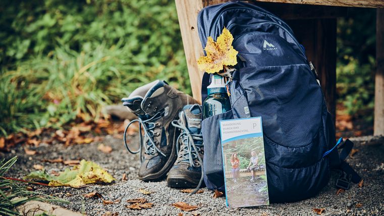 Ein Paar Wanderschuhe lehnt an einer hölzernen Bank, daneben ein blauer Rucksack und ein Wanderflyer.