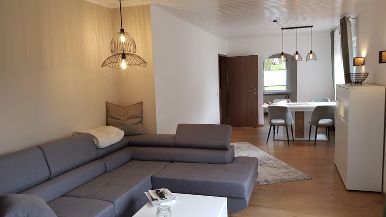 Ein modernes Wohnzimmer mit einem grauen Sofa und einem minimalistischen Tisch. Im Hintergrund befindet sich ein Essbereich mit Stühlen und einem Fenster.