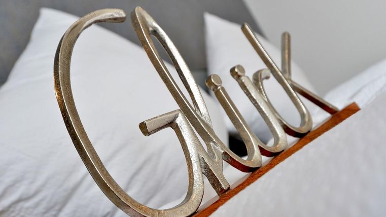 Ein dekoratives Wort "Glück" aus Metall steht auf einem Bett. Im Hintergrund sind zwei weiße Kissen sichtbar.