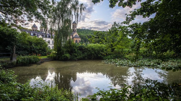 Ein malerischer Teich, umgeben von üppigem Grün und spärlichem Unterholz. Im Hintergrund ist ein Gebäude mit Türmen und Zypressen sichtbar, reflektiert im Wasser.