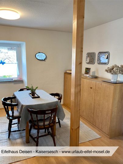 Ein gemütlicher Essbereich mit einem Holztisch und Stühlen. An der Wand stehen Regale mit Dekorationen und einer kleinen Blumenvase.