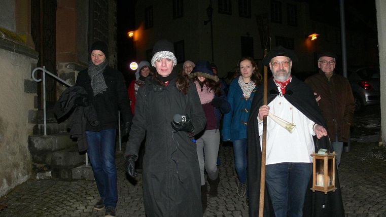 Eine Gruppe von Menschen geht nachts durch eine kopfsteingepflasterte Straße. Die Person vorne trägt eine Laterne und ist in traditioneller Kleidung gekleidet.