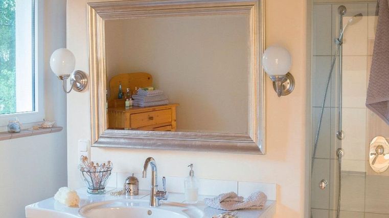 Une salle de bain lumineuse avec un miroir élégant et un lavabo. En arrière-plan, on peut voir une douche, et il y a des éléments décoratifs sur le rebord.