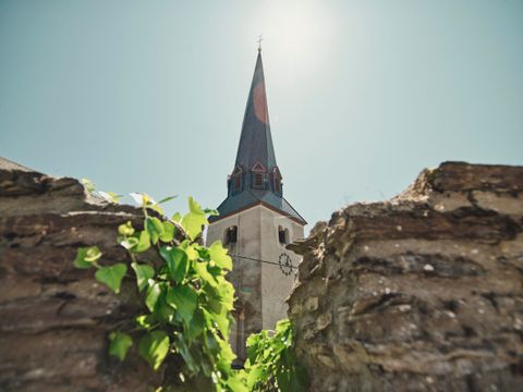 Ein Blick auf den Kirchturm einer Kirche, umgeben von einer alten Mauer und grünen Pflanzen. Der Himmel ist klar und die Sonne scheint.