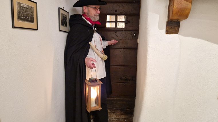 Een man in historische kleding staat in een gang en houdt een lantaarn vast. Achter hem is een houten deur en aan de muur hangen schilderijen.