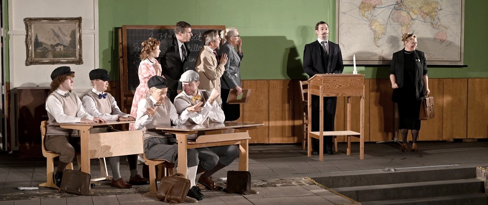 Klassenzimmerszene aus der Auff&uuml;hrung des Theaterverein Kaisersesch des St&uuml;cks &bdquo;Die Feuerzangenbowle&ldquo;: Mehrere Darsteller in historischen Schuluniformen mit Schieberm&uuml;tzen sitzen an alten Holzb&auml;nken, w&auml;hrend zwei Lehrerfiguren und weitere Erwachsene vor einer Tafel stehen. Rechts spricht ein Lehrer mit Schnurrbart am Pult, neben ihm eine streng wirkende Lehrerin mit Aktentasche. Im Hintergrund h&auml;ngt eine gro&szlig;e Weltkarte, die B&uuml;hne ist als nostalgisches Klassenzimmer gestaltet.
