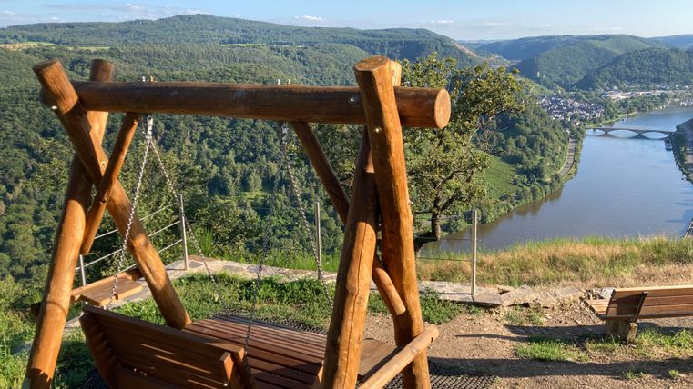 Ein Holzschaukel auf einem Aussichtspunkt mit Blick auf einen Fluss und die umgebenden Hügel. Die Landschaft ist grün und der Himmel ist klar.