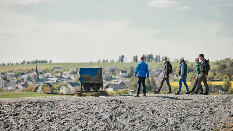 Een groep van vijf personen loopt over een heuvel met uitzicht op een dorp. Op de voorgrond staat een blauwe kar.