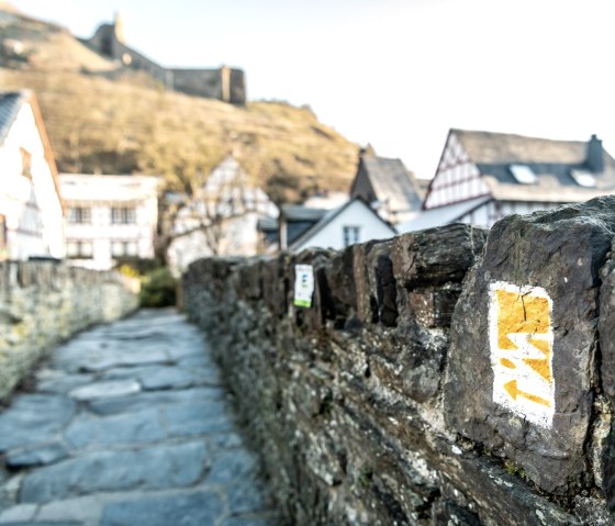 Wanderweg-Markierung auf einer Steinmauer, im Hintergrund Fachwerkhäuser und eine Burg auf einem Hügel., © Eifel Tourismus GmbH, D. Ketz