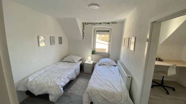 Ein helles Schlafzimmer mit zwei Einzelbetten und einem Fenster. An der Wand sind Bilderrahmen und ein kleiner Schreibtisch mit einem Stuhl zu sehen.