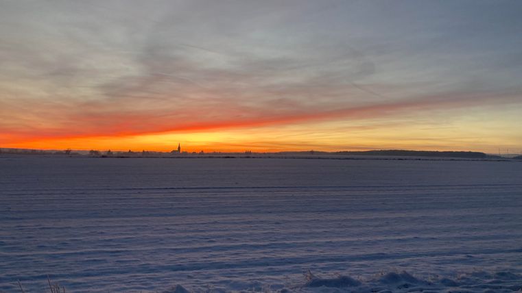 Ein Winterlandschaft mit einer schneebedeckten Fläche und einem farbenfrohen Sonnenuntergang. Der Himmel zeigt sanfte Orange- und Blautöne.