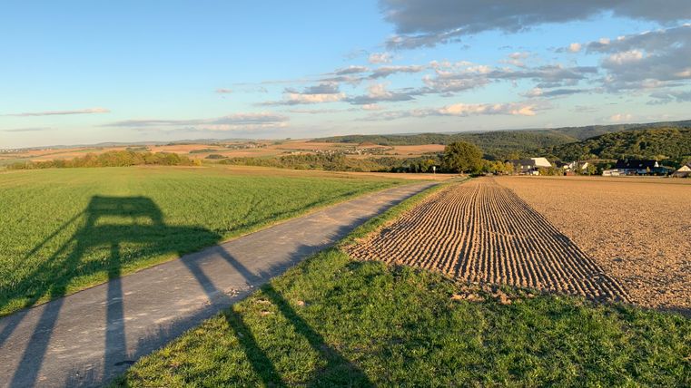 A quiet landscape with a hiking trail that winds through lush meadows and freshly tilled fields. The sky is clear with some clouds, and nature sparkles in soft colors.