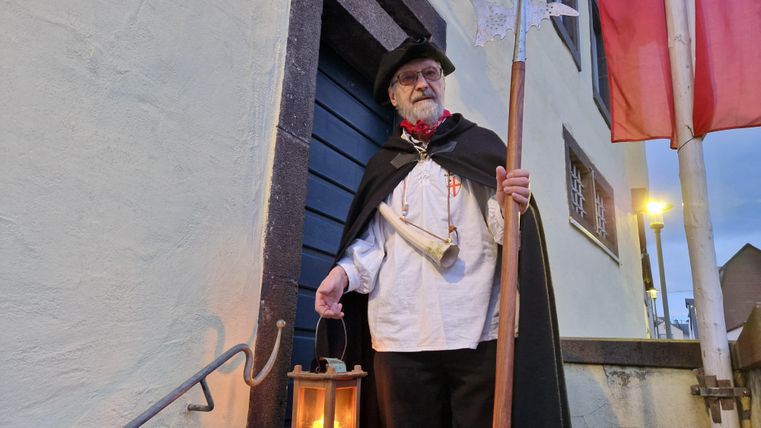 Ein Mann in traditioneller Kleidung steht vor einer Tür, während er eine Laterne und einen Stock hält. Im Hintergrund sind fahnen und eine sanfte Abendbeleuchtung zu sehen.