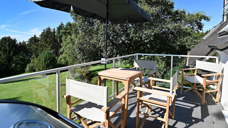 Ein schöner Balkon mit einem Tisch und Stühlen, umgeben von Bäumen. Die Terrasse hat einen Sonnenschirm und bietet einen Blick ins Grüne.
