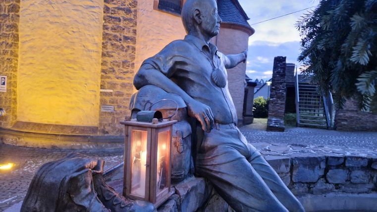 Een standbeeld van een man die ontspannen zit en naar iets wijst. Naast hem staat een lamp die zachtjes gloeit.