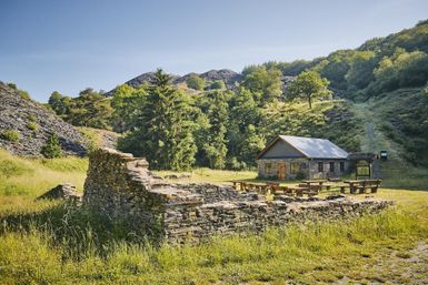 An old stone ruin stands in a green landscape. In the background, there is a modern, typically Scottish wooden building.