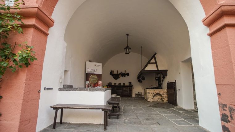 Ein traditioneller Innenraum mit einem großen gewölbten Eingang. Auf der linken Seite sind Tische und eine Küche sichtbar.