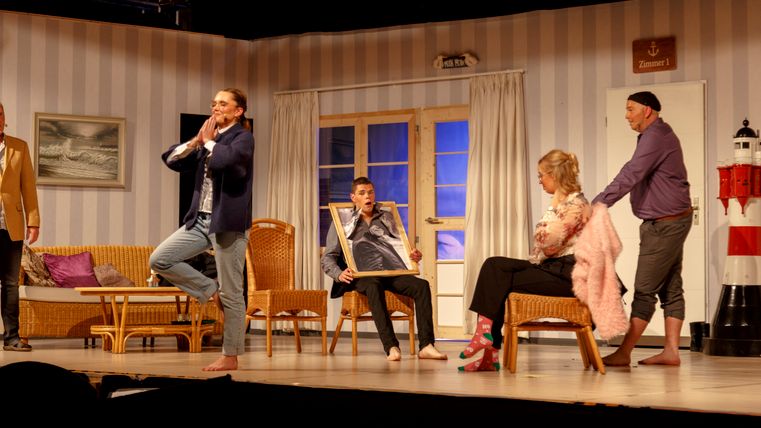 Theateraufführung auf einer Wohnzimmerbühne: Mehrere Darstellende in einer komödiantischen Szene, mit Korbmöbeln, Leuchtturm-Dekoration und einem großen gerahmten Bild als Requisite.
