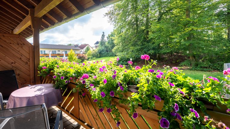 Ein schöner Balkon mit bunten Blumen und einem Tisch. Im Hintergrund sieht man Bäume und ein Haus.